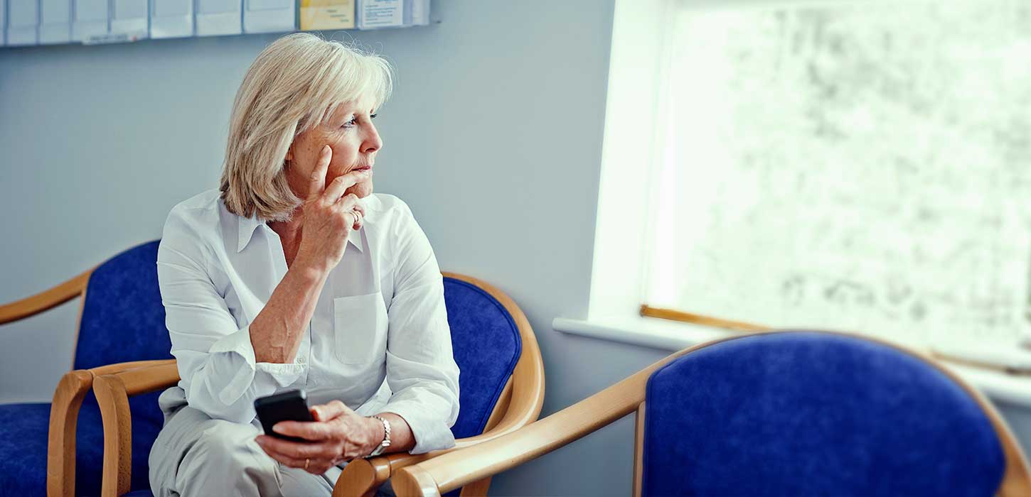 A photo of an elderly woman, sitting alone in a hospital waiting room. She looks worried as she stares out the window.